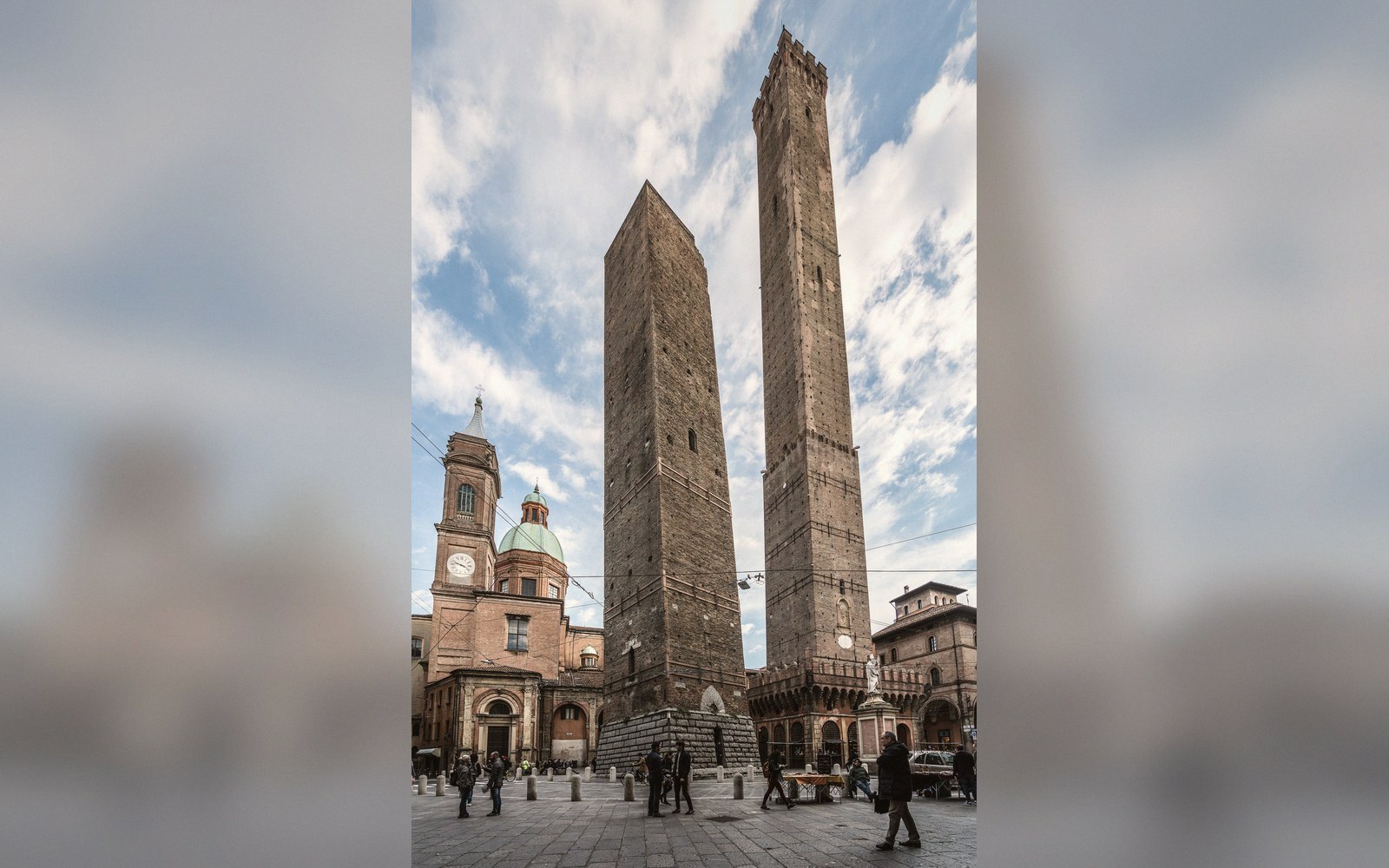 Two leaning medieval brick towers rise above a cobbled square with pedestrians below