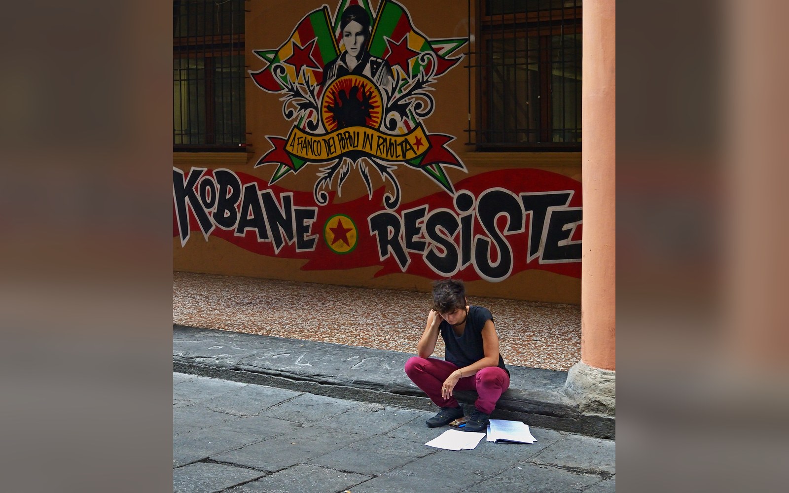 A person crouches on a sidewalk beneath a "Kobane Resiste" mural on a wall