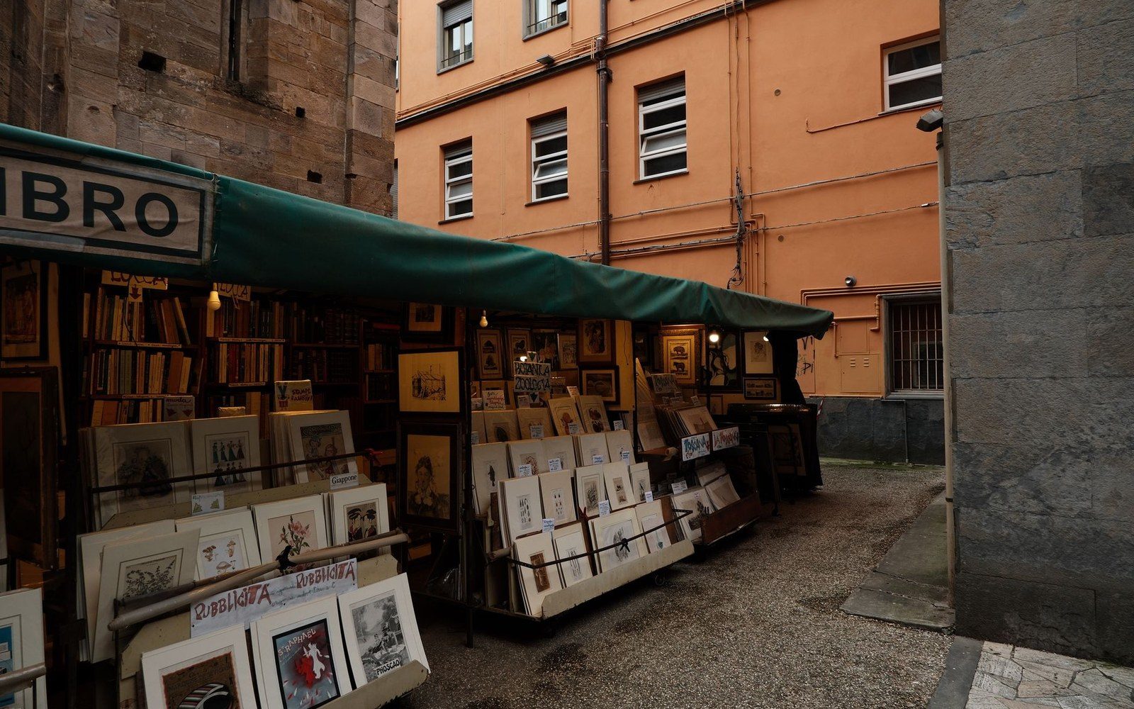 Outdoor bookstall with green awning displaying prints and books beside a pink building