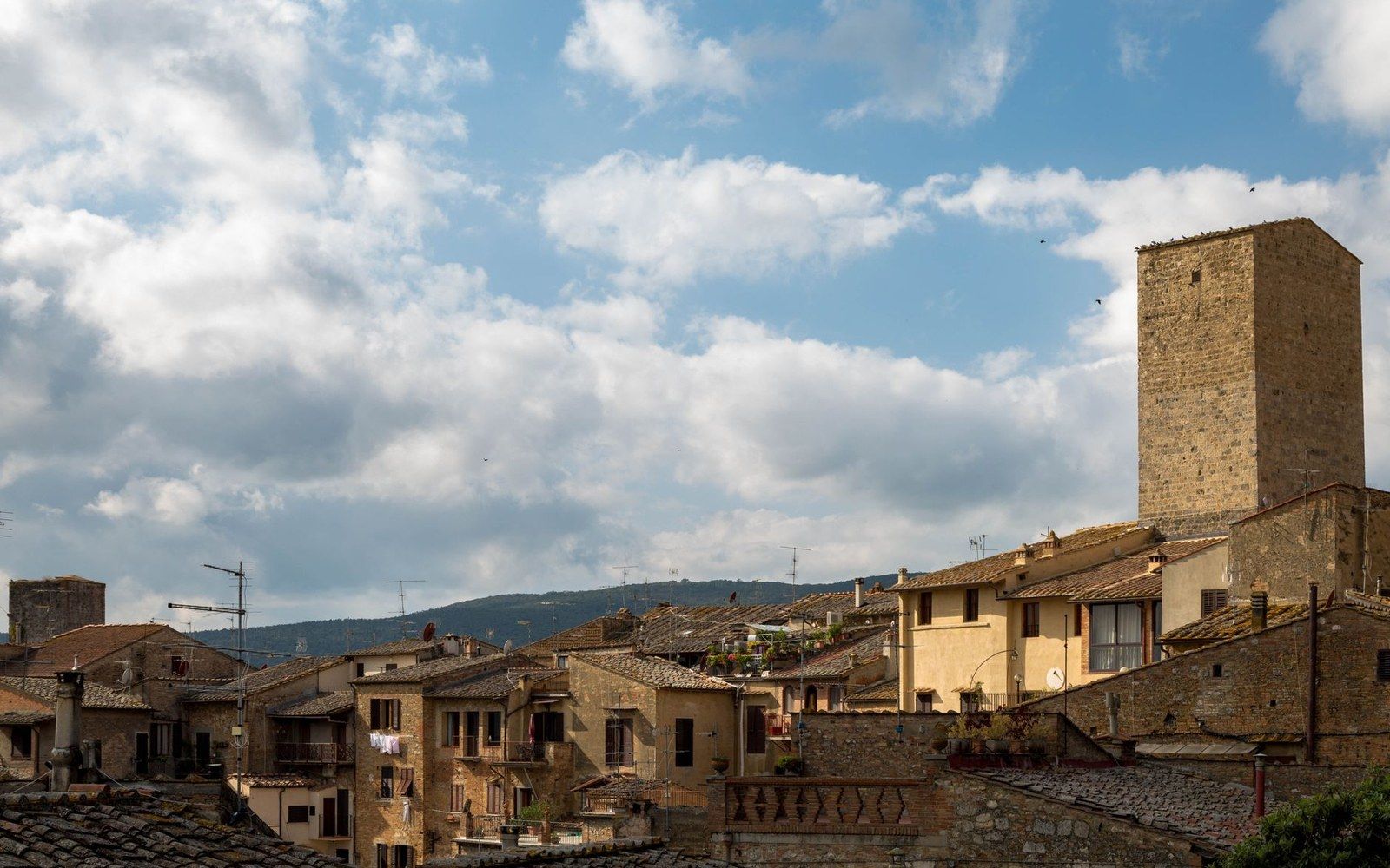A stone medieval tower rises above a hillside town's terracotta rooftops under a partly cloudy sky